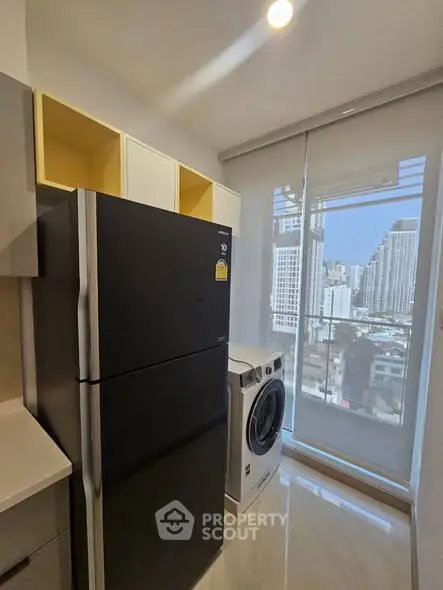 Modern kitchen with sleek black fridge and washing machine, offering city views through large window.