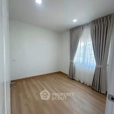 Spacious empty room with wooden flooring and large window with curtains.