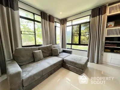 Spacious and sunlit living room with large windows offering a serene garden view, complemented by a plush grey sectional sofa and elegant draperies.