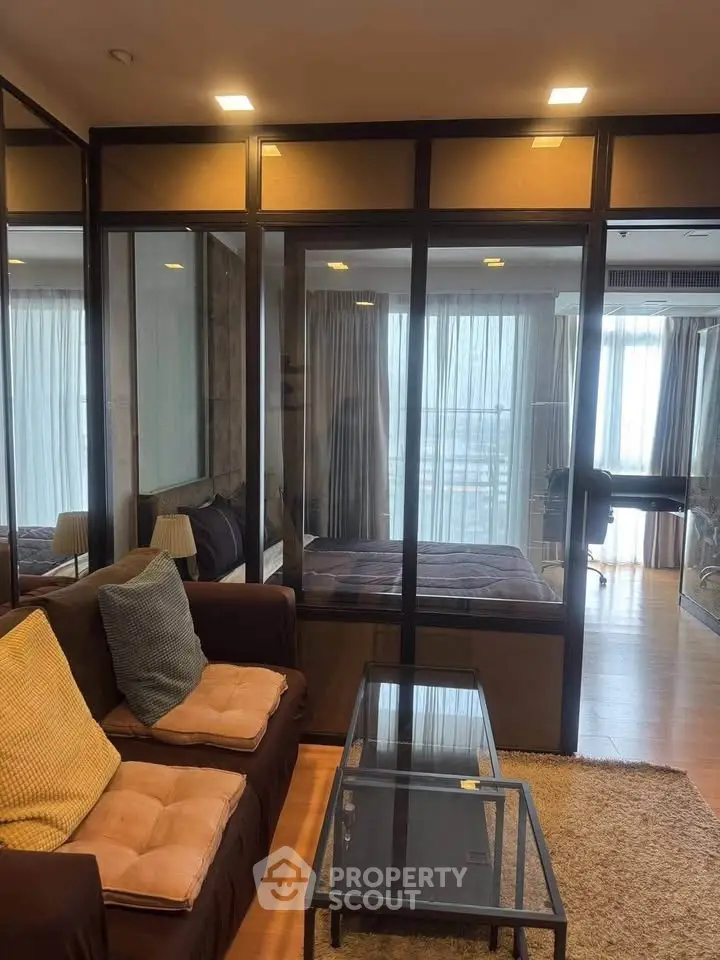 Modern apartment with glass partition separating living room and bedroom, featuring cozy seating and natural light.