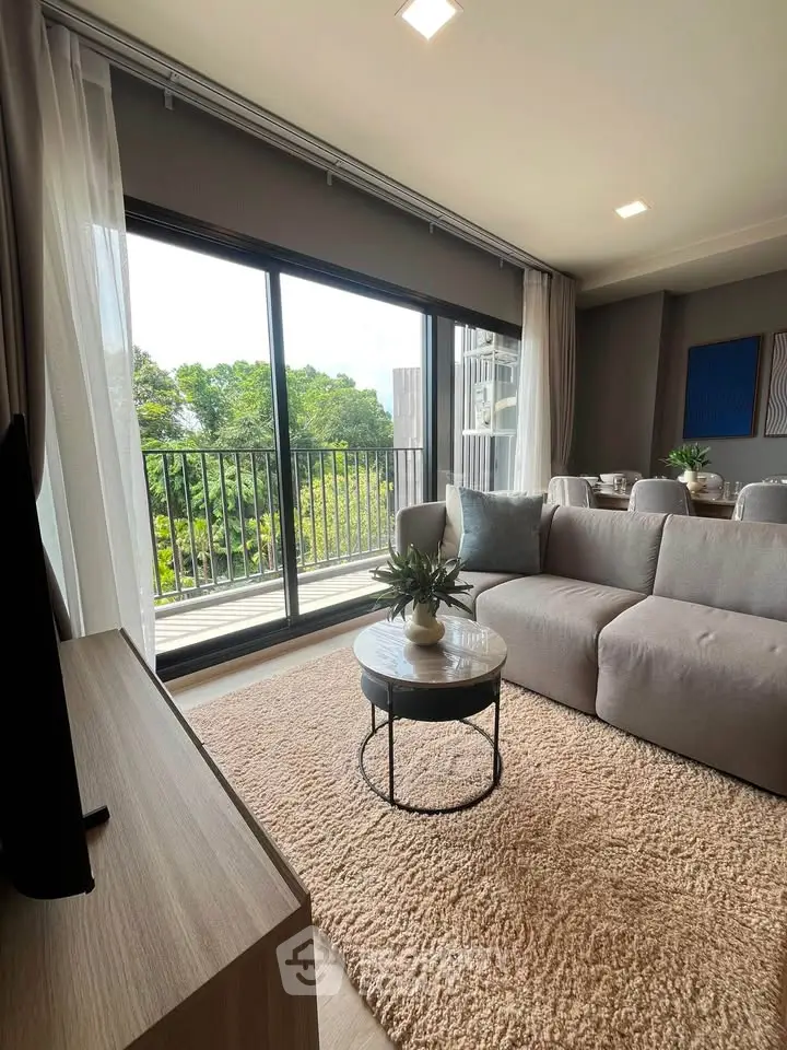 Modern living room with large windows and balcony view, featuring stylish furniture and lush greenery outside.