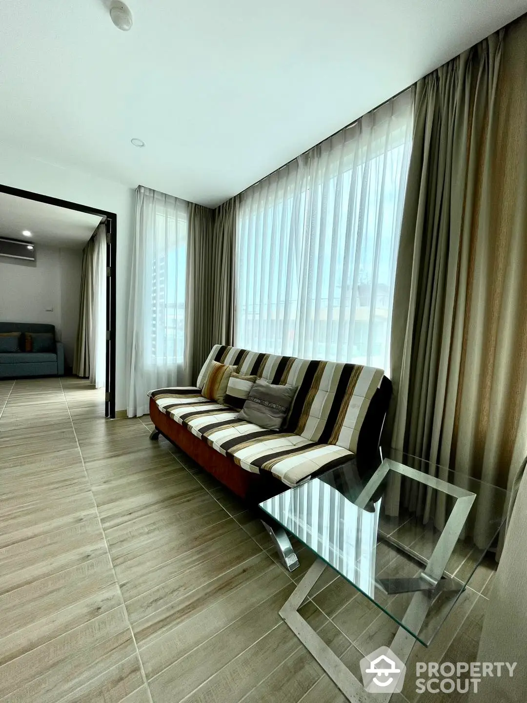 Spacious and modern living room with large windows draped in sheer curtains, elegant glass coffee table, and a plush striped sofa, leading to an adjoining room.