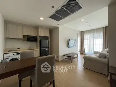 Modern living room with open kitchen, featuring sleek furniture and natural light from large windows.