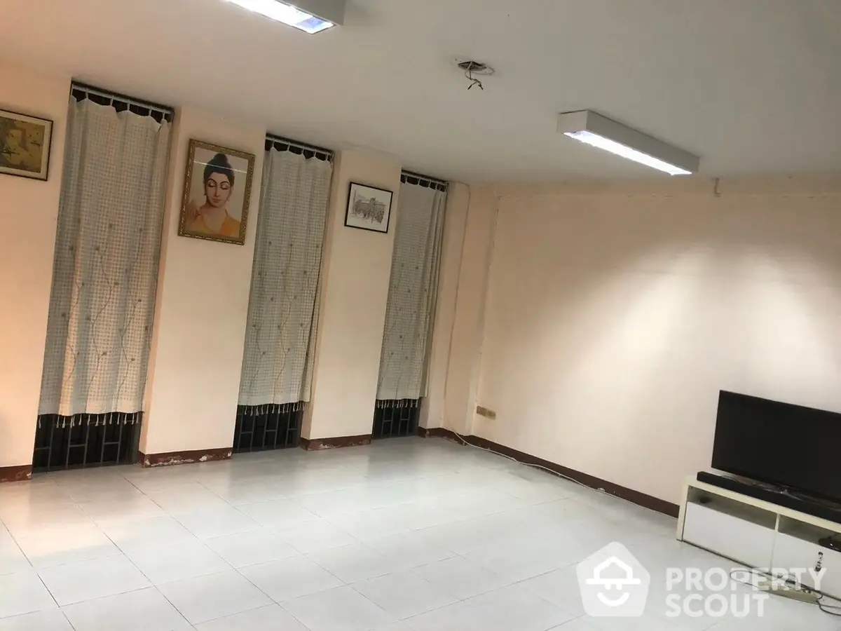 Spacious and well-lit living room with pristine white tiles, ample natural light from large windows with patterned curtains, and a modern flat-screen TV.