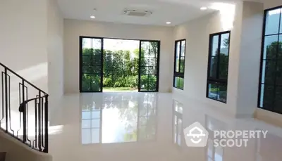 Spacious and bright living room with glossy white tiles, large windows offering a serene garden view, and a modern staircase leading to the upper level.