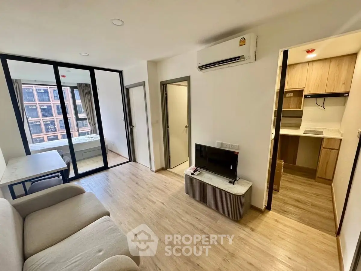 Modern living room with open kitchen and natural light, featuring sleek furniture and wooden flooring.