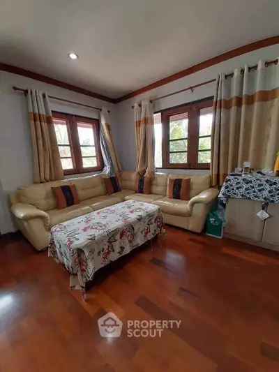 Cozy living room with wooden flooring and large windows, featuring a comfortable beige sofa and floral tablecloth.