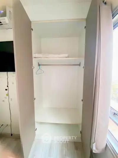 Spacious white wardrobe with hanging rod and shelf in bright room with window view.