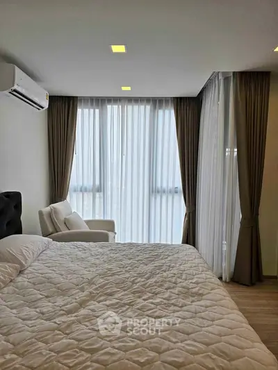 Spacious bedroom with large windows and elegant curtains, featuring a cozy armchair and modern air conditioning.