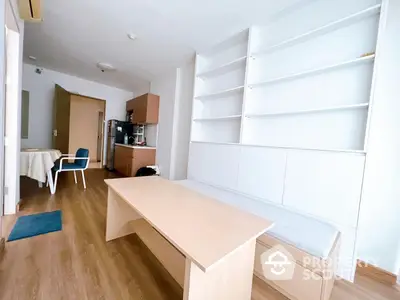 Spacious and well-lit living room with modern wooden flooring, large white shelving unit, and minimalist furniture, leading to a cozy kitchen area.