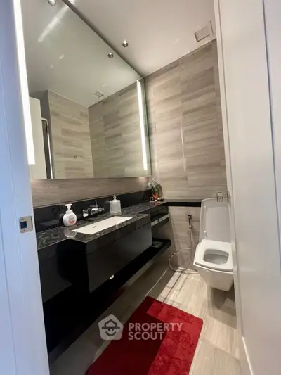 Modern bathroom with sleek design, featuring a large mirror, elegant vanity with dark countertop, and stylish tiling throughout, complemented by a red accent rug.