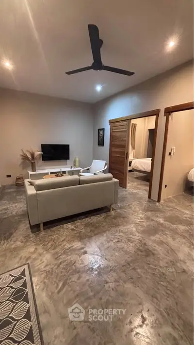 Modern living room with concrete flooring, ceiling fan, and cozy seating area leading to a bedroom.