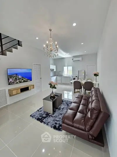 Modern living room with elegant chandelier and open kitchen layout, featuring sleek furniture and a large TV.