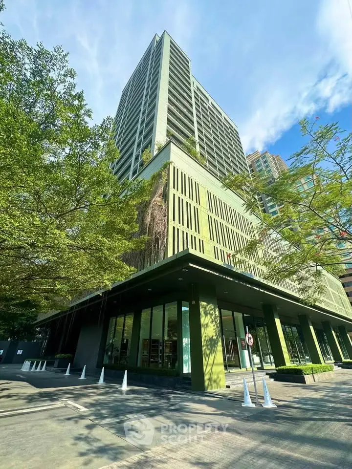 Modern green building exterior with lush trees and clear blue sky, showcasing urban architecture.