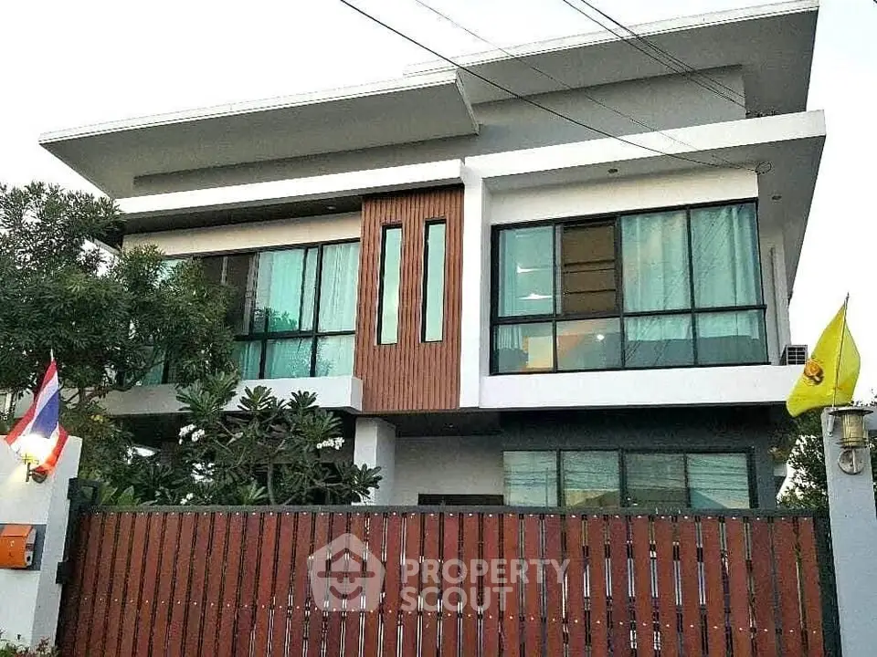 Modern two-story house with sleek design and large windows, surrounded by lush greenery and a wooden fence.
