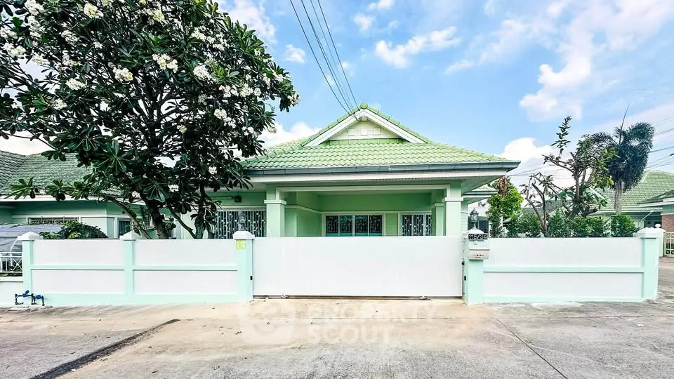 Charming single-story green house with lush garden and secure white fence in tranquil neighborhood.