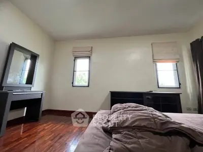 Spacious bedroom with wooden flooring and natural light from two windows, featuring a bed and a dressing table.