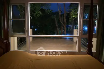 Spacious bedroom with large window offering serene night view of trees and balcony.