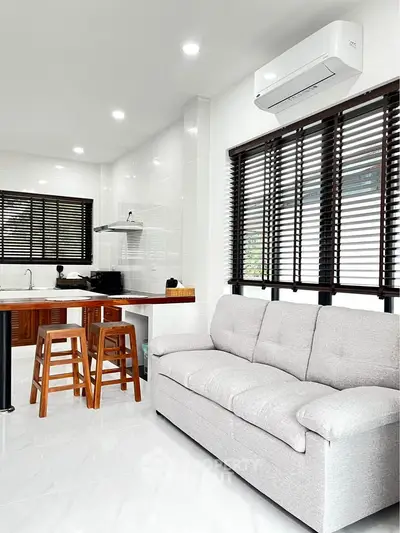 Modern living room with open kitchen, featuring sleek sofa and bar stools under natural light.