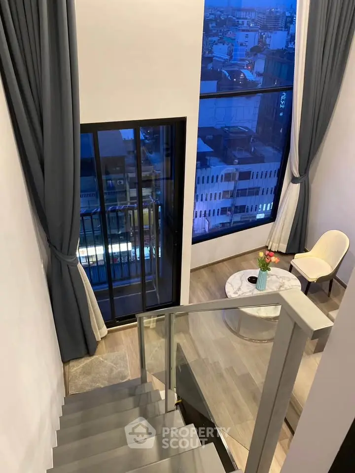 Modern duplex interior with large windows and city view, featuring elegant staircase and cozy seating area.
