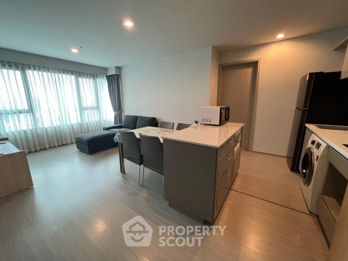 Modern open-plan living room with kitchen, featuring sleek furniture and natural light from large windows.