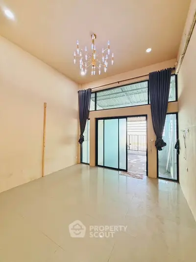 Spacious living room with high ceiling and elegant chandelier, featuring large windows and balcony access.