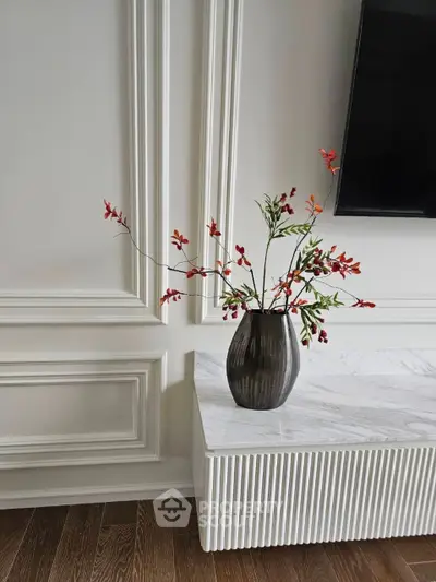 Elegant interior with classic white wall molding, a sleek flat-screen TV, and a stylish vase with vibrant flowers on a marble table, showcasing a modern yet timeless design.