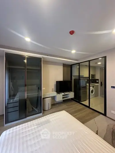 Modern bedroom with glass wardrobe and view into kitchen with washing machine.