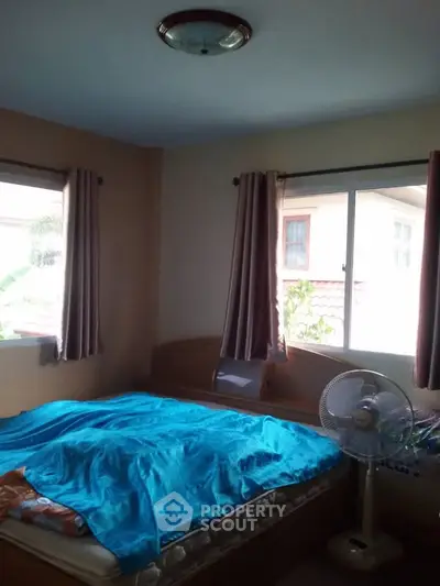 Cozy bedroom with large windows and natural light, featuring a comfortable bed and standing fan.