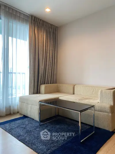 Modern living room with stylish beige sofa and glass coffee table, complemented by a blue rug and floor-to-ceiling window.