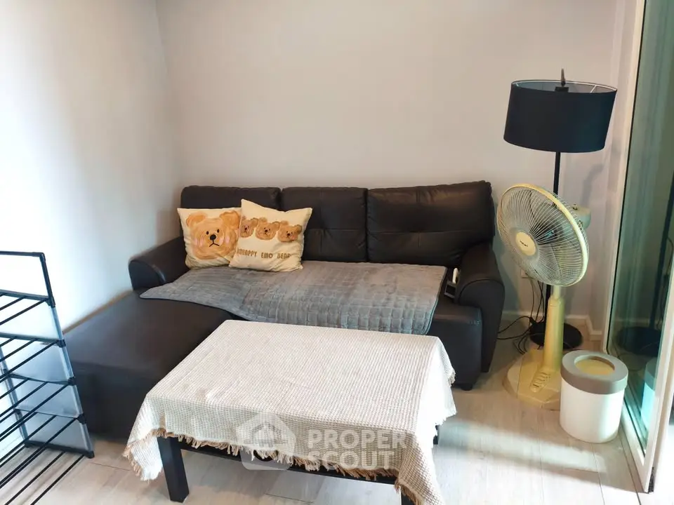 Cozy living room with a black leather sofa, decorative pillows, and a standing fan, perfect for relaxation.