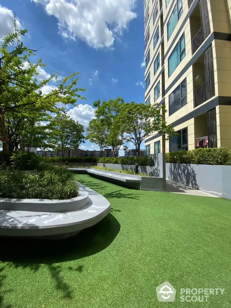 Modern apartment building with lush green garden and seating area under blue sky.