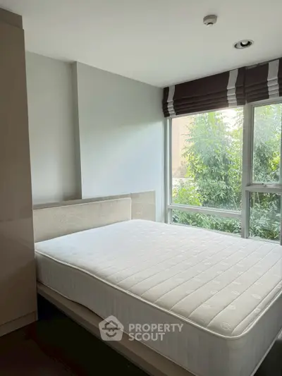 Bright bedroom with large window and comfortable mattress, offering a serene view of lush greenery.