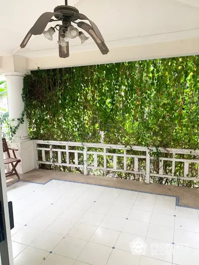 Spacious covered balcony with lush greenery wall, ceiling fan, and tiled flooring, perfect for outdoor relaxation and entertainment in a serene setting.