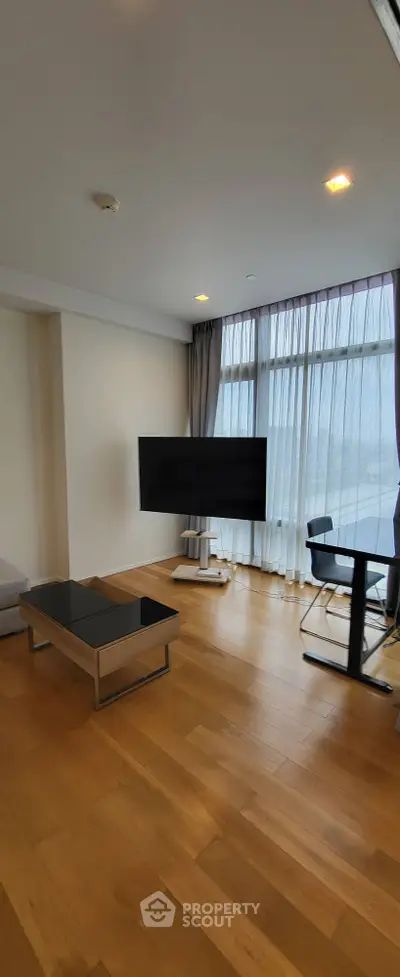 Modern living room with sleek TV and elegant furniture, featuring floor-to-ceiling windows and wooden flooring.