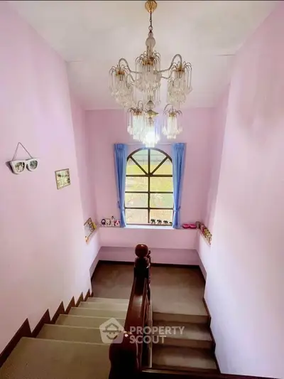 Charming staircase with elegant chandelier and pastel pink walls, featuring a large arched window with blue curtains.