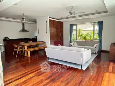 Spacious living room with open kitchen and wooden flooring, featuring modern furniture and large windows.