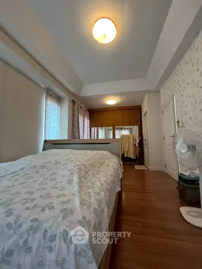 Cozy bedroom with wooden flooring and floral wallpaper, featuring a comfortable bed and ample natural light.