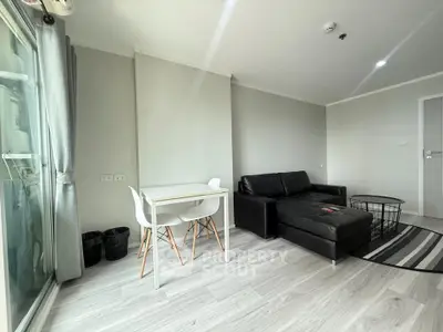 Modern living room with sleek black sofa and dining area, featuring large windows and light wood flooring.