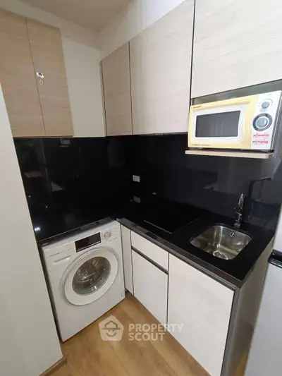 Modern compact kitchen with washing machine and microwave, sleek black countertops, and wooden cabinets.