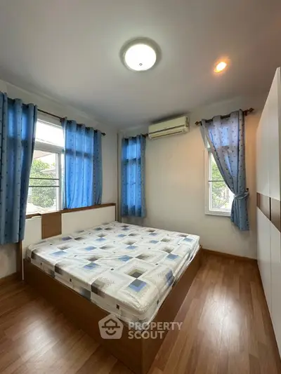 Cozy bedroom with wooden flooring and blue curtains, featuring a large window for natural light.