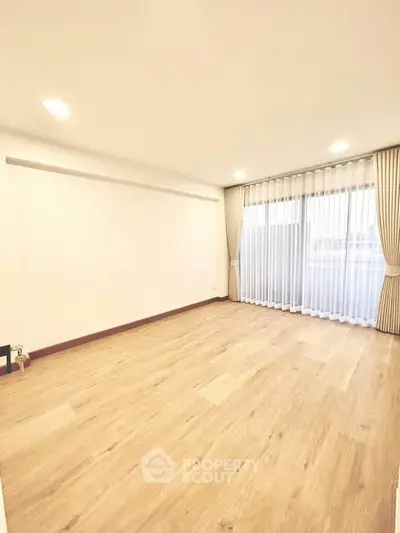 Spacious empty room with wooden flooring and large window with curtains