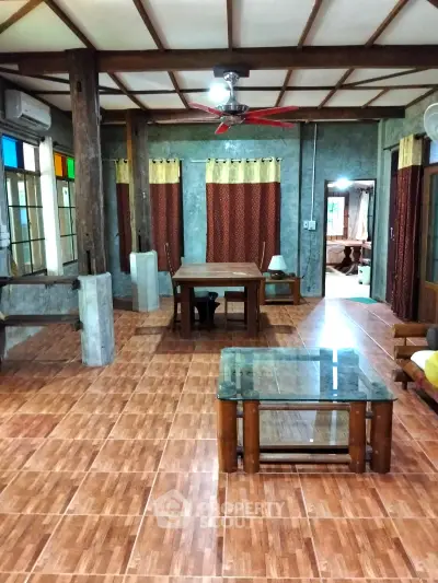 Spacious living room with rustic wooden decor and tiled flooring, featuring large windows and ceiling fan.
