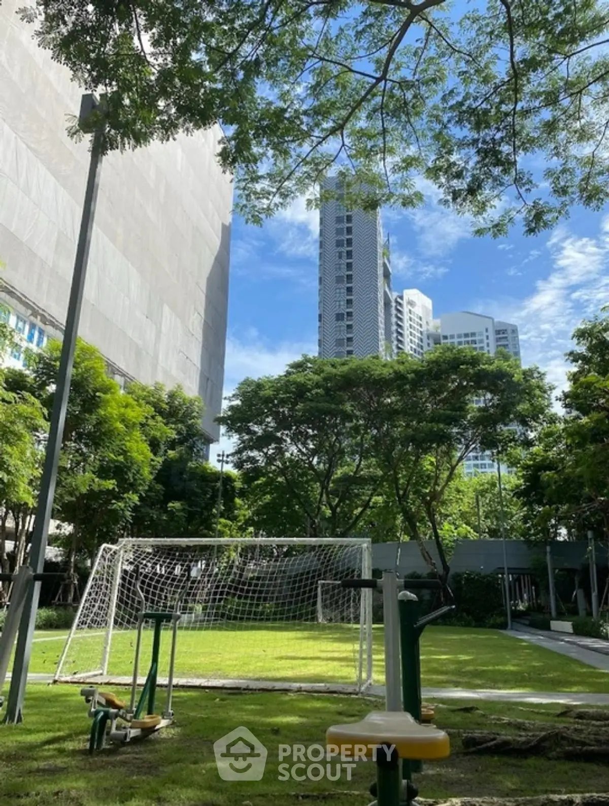 Urban park with exercise equipment and soccer goal, surrounded by skyscrapers and lush greenery.