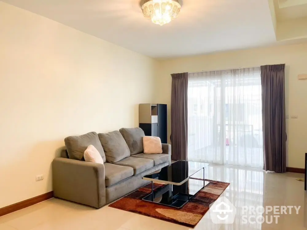 Spacious and well-lit living room with elegant furnishings, large windows draped with maroon curtains, and a cozy grey sofa set on a warm-toned rug.