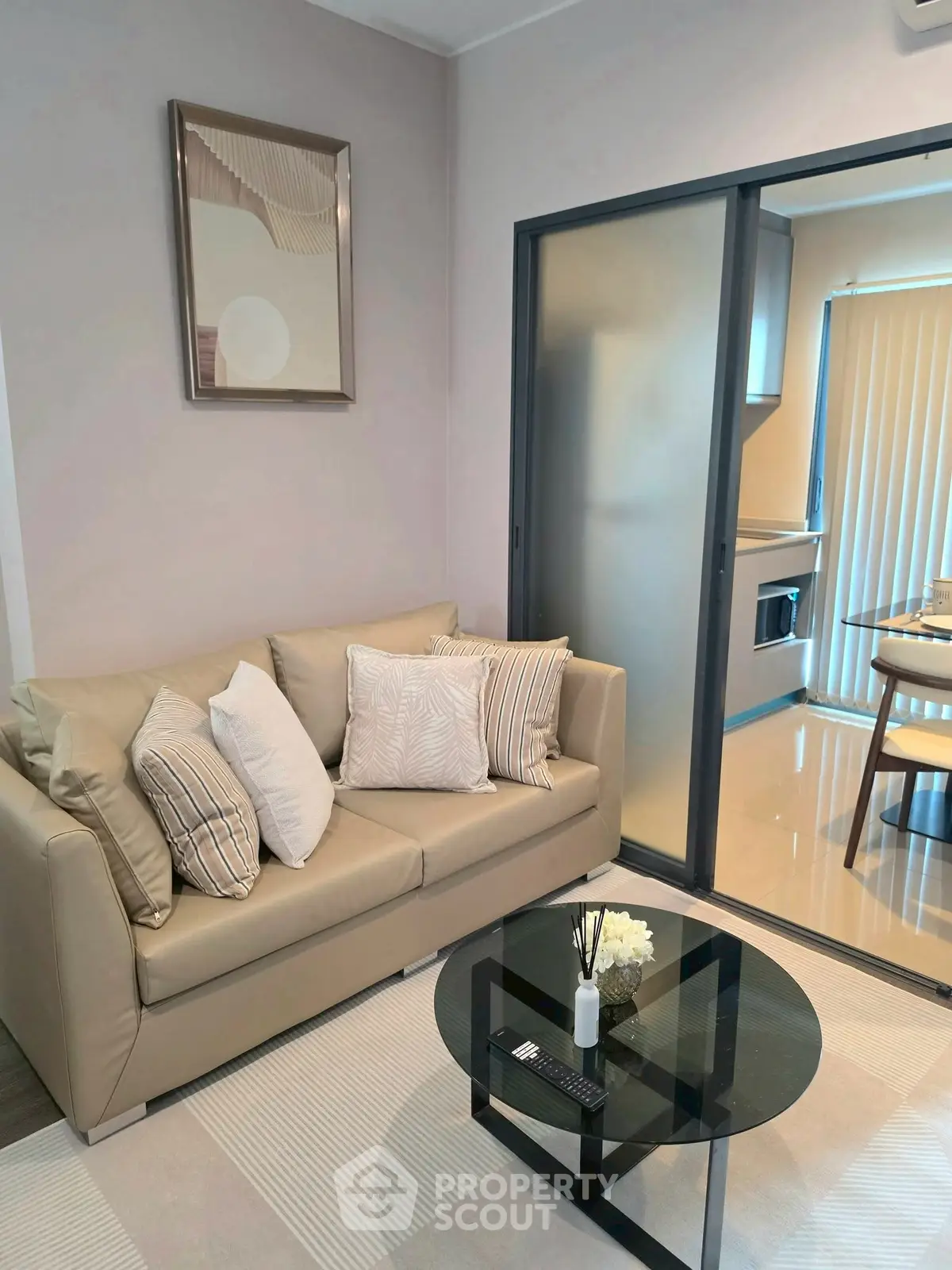 Modern living room with beige sofa and glass coffee table, adjacent to a sleek kitchen with sliding doors.