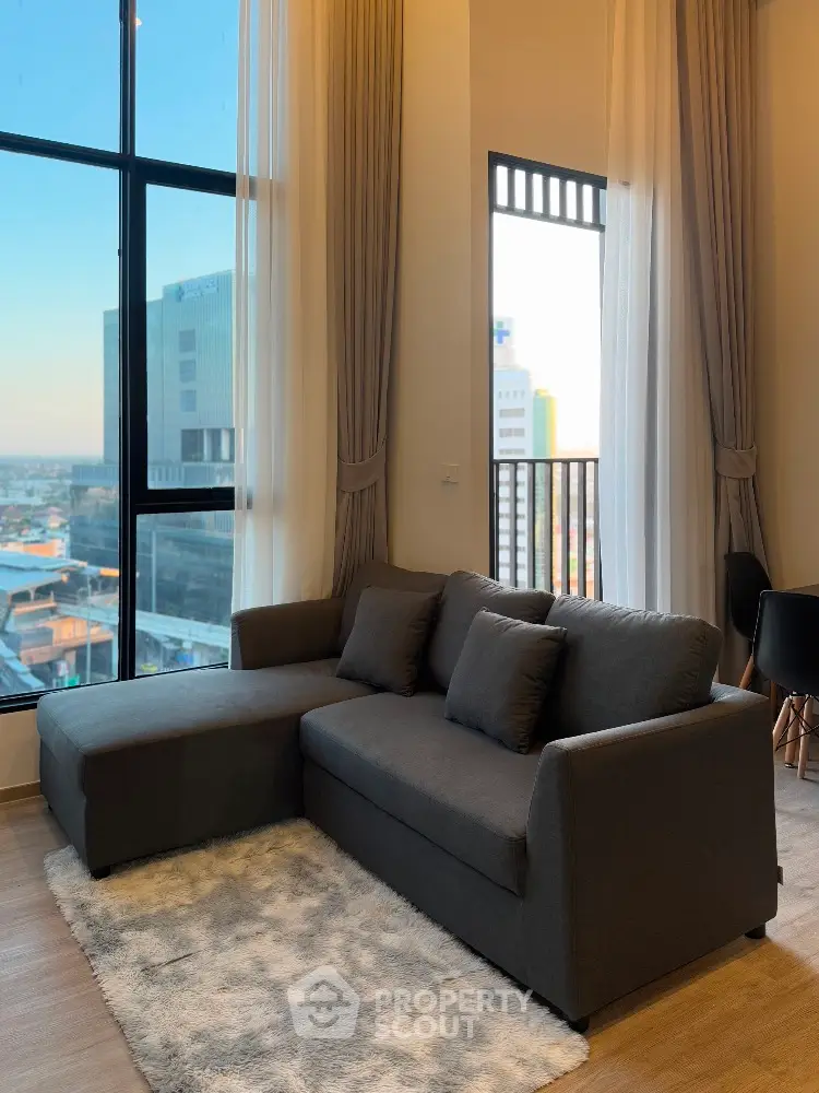 Modern living room with large windows and city view, featuring a stylish gray sofa and cozy rug.