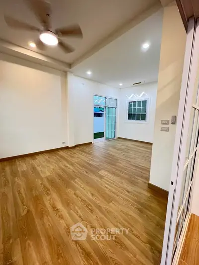 Spacious living room with wooden flooring and ceiling fan, bright natural light.