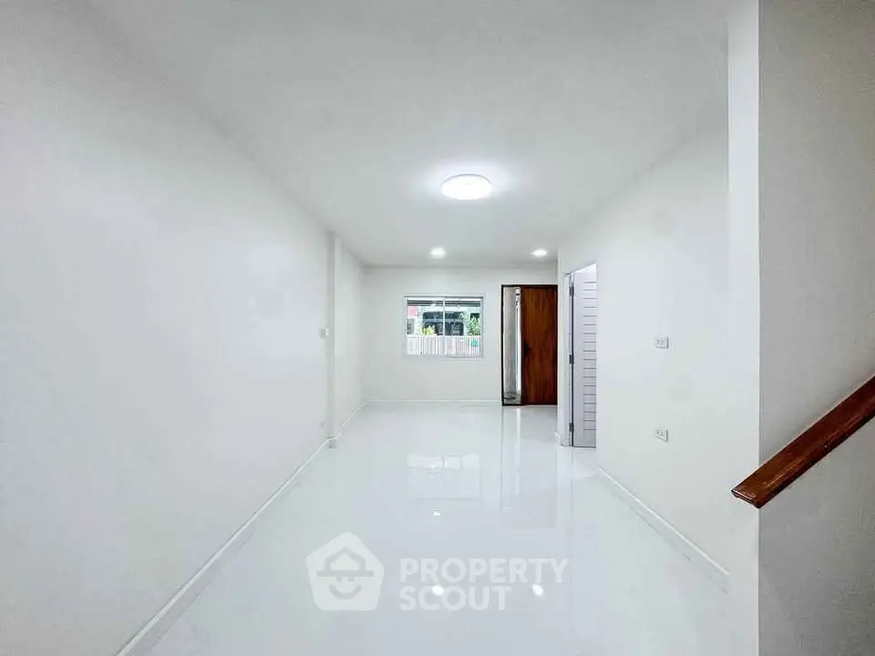Spacious minimalist living room with white walls and glossy floor, featuring a large window and wooden door.