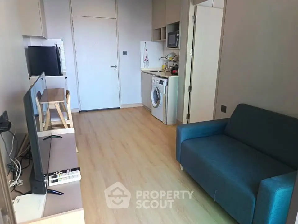 Modern compact living room with integrated kitchen and washing machine, featuring sleek furniture and wooden flooring.